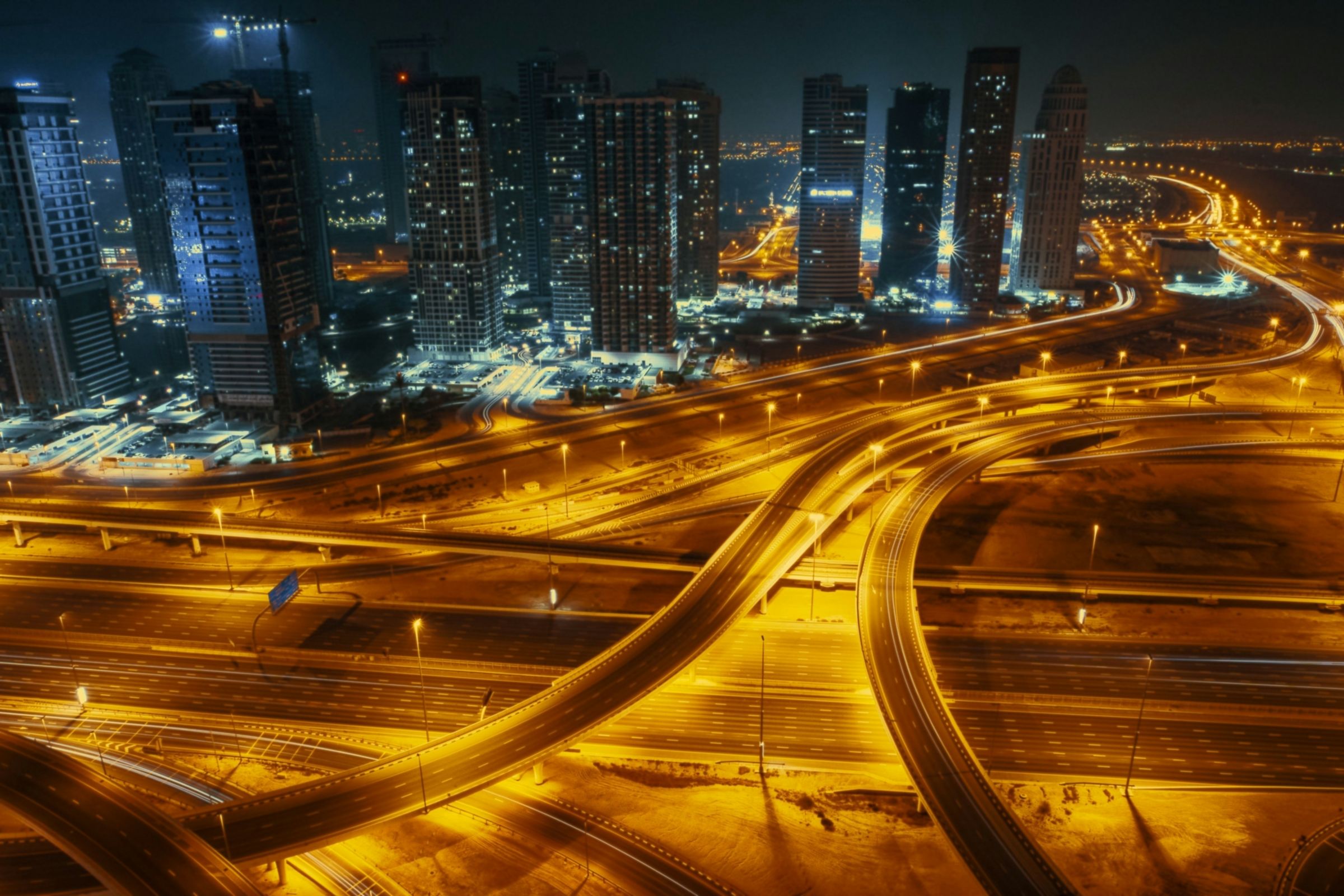 Aerial view of Dubai at night, with a multi-level highway interchange curving beneath a line of lit towers under a dark sky.