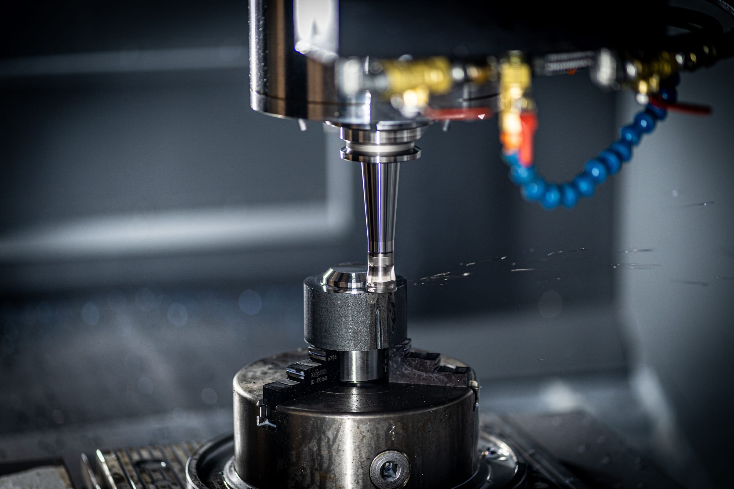 A milling spindle lowered onto a metal workpiece clamped in a chuck, with a blue coolant line curving in from the right.