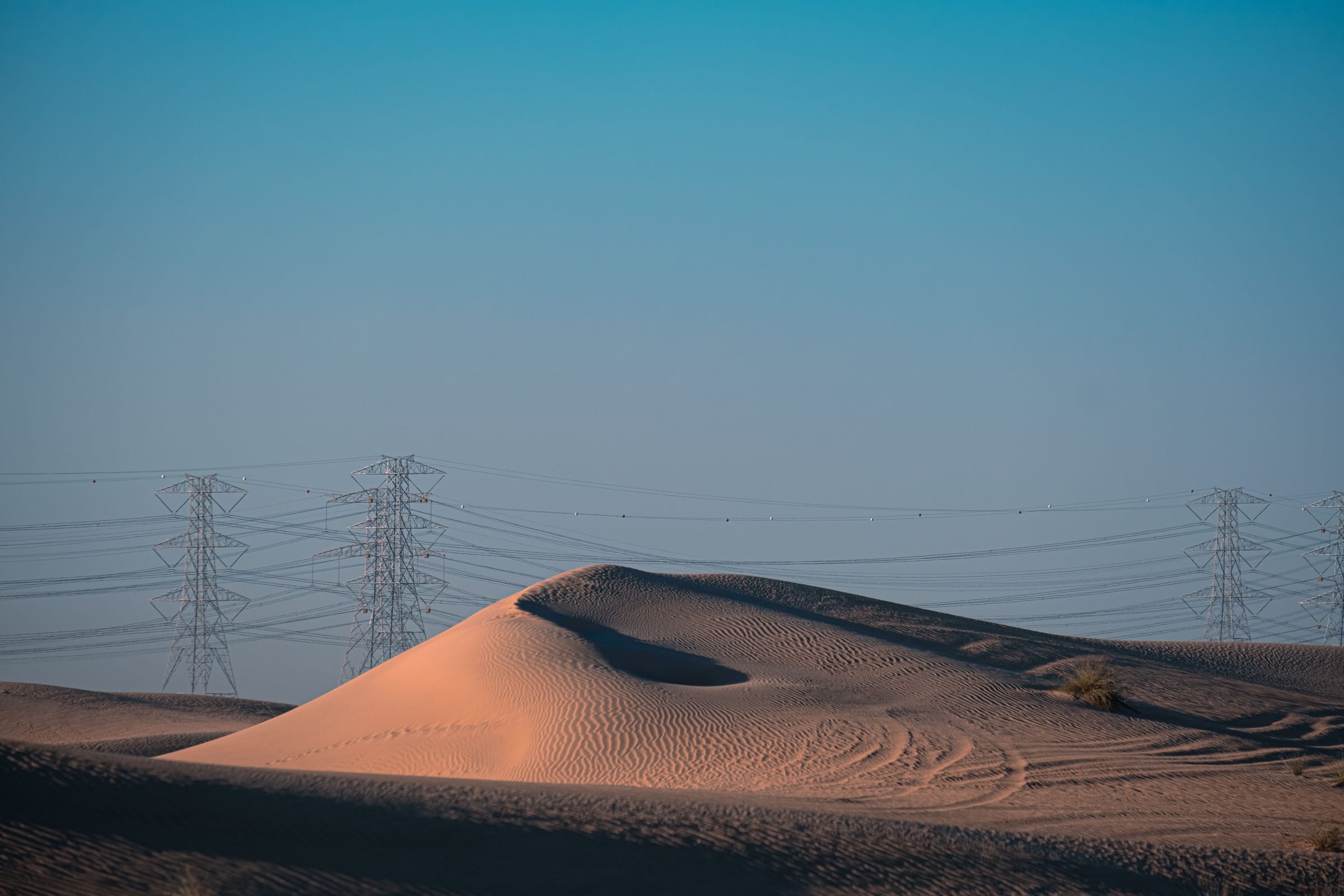 A sand dune in the foreground with a line of electricity transmission towers behind it under a clear blue sky.