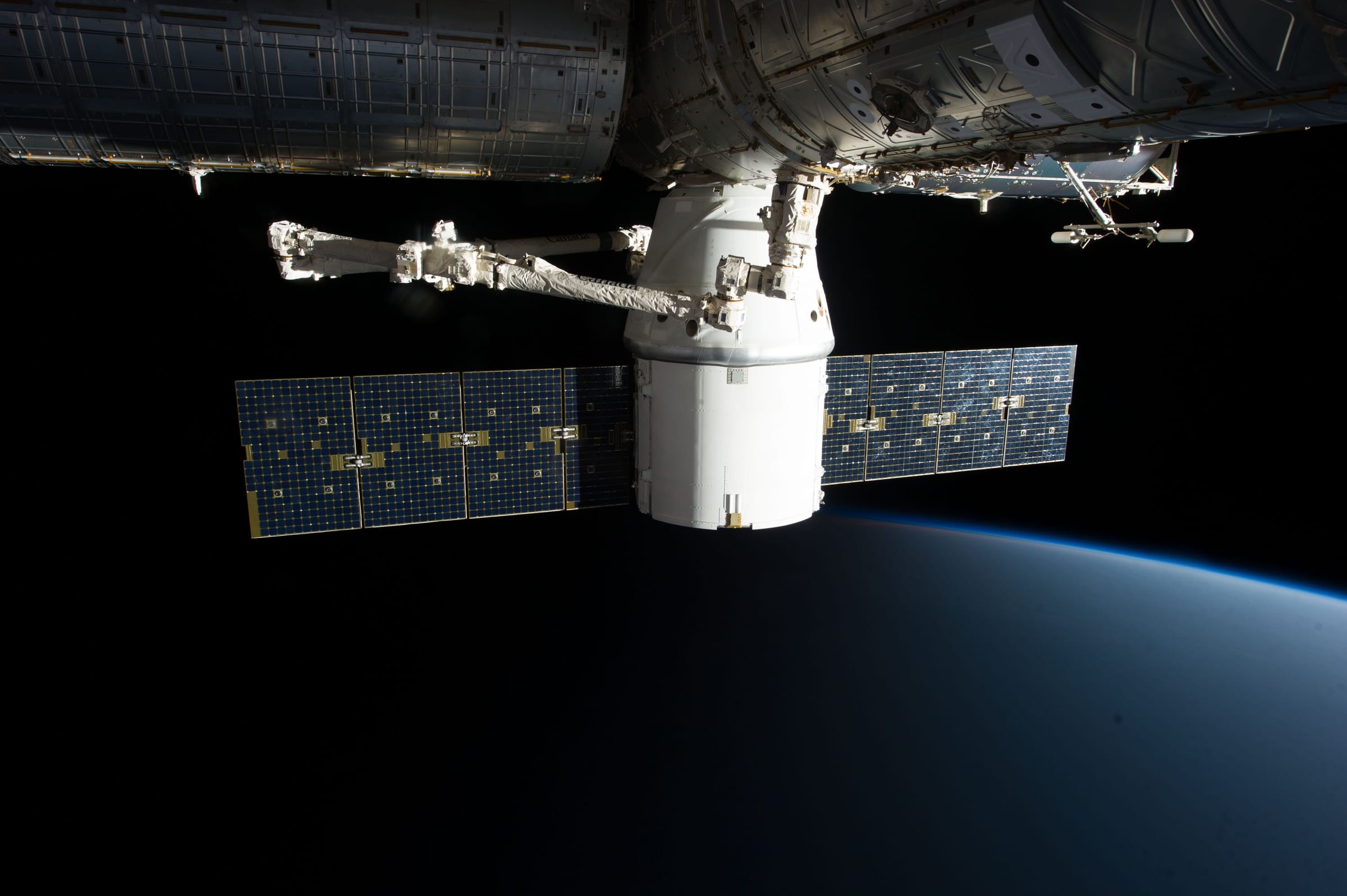 A cargo spacecraft with extended solar panels held by a robotic arm below the International Space Station, with the curved edge of Earth beneath.