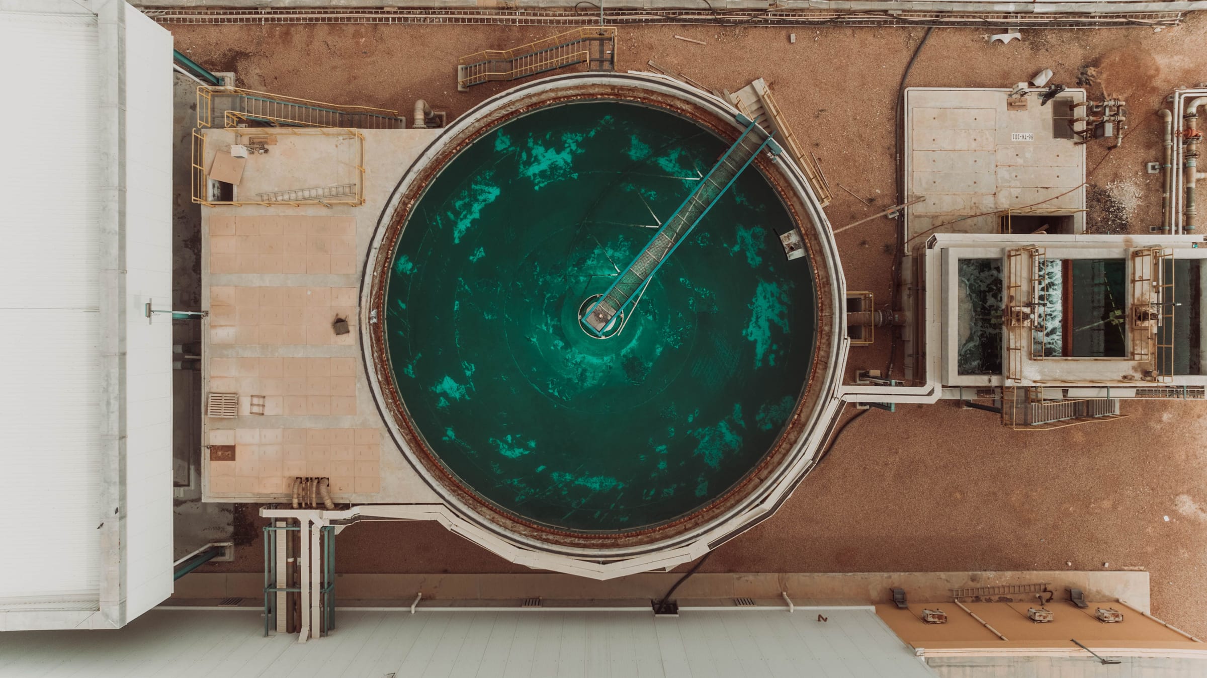 Overhead view of a circular water treatment tank filled with deep green water, its rotating bridge reaching to the centre, surrounded by pale concrete and sand.