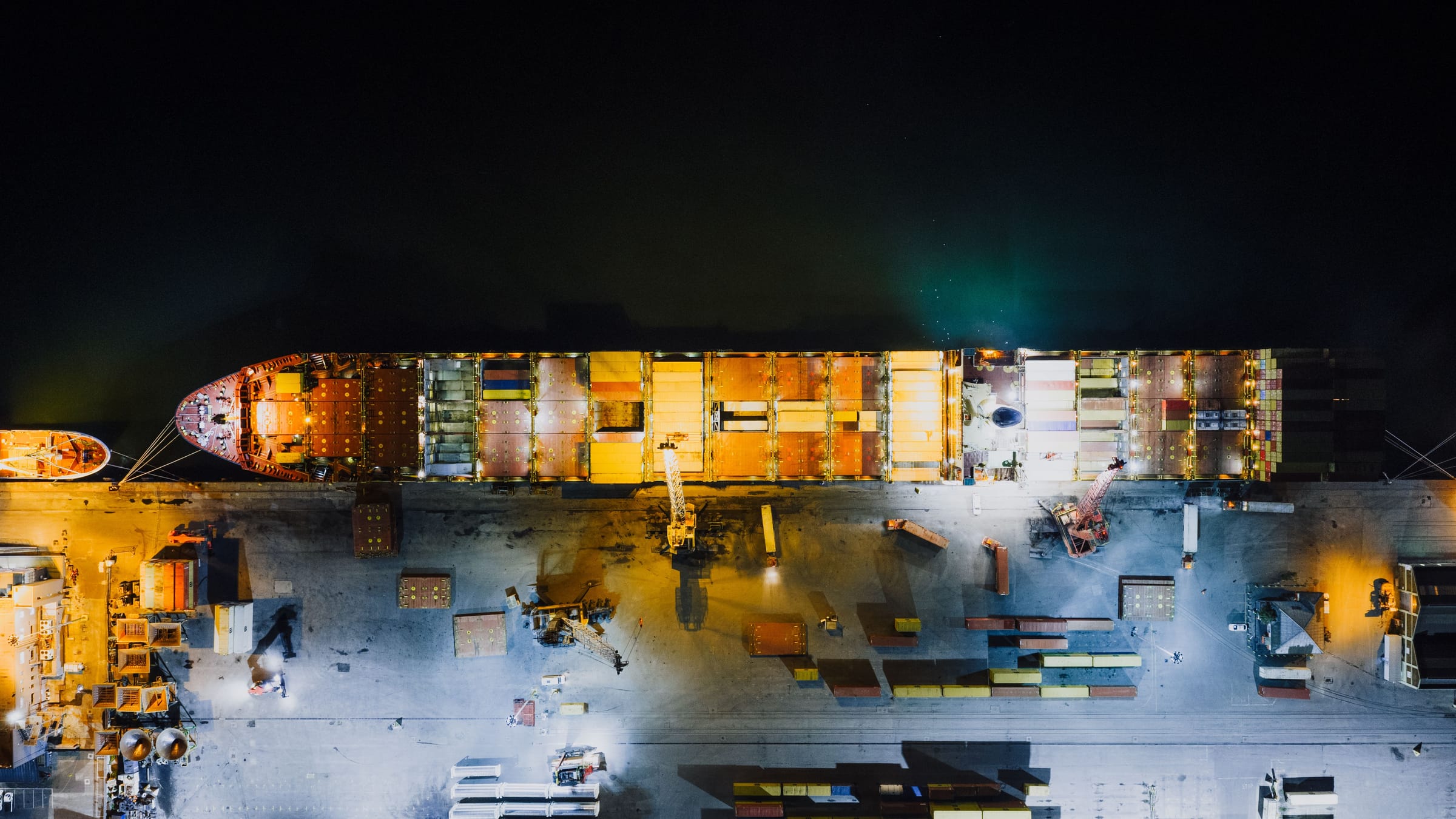 Overhead night view of a container ship moored at a floodlit quay, with dark water above.