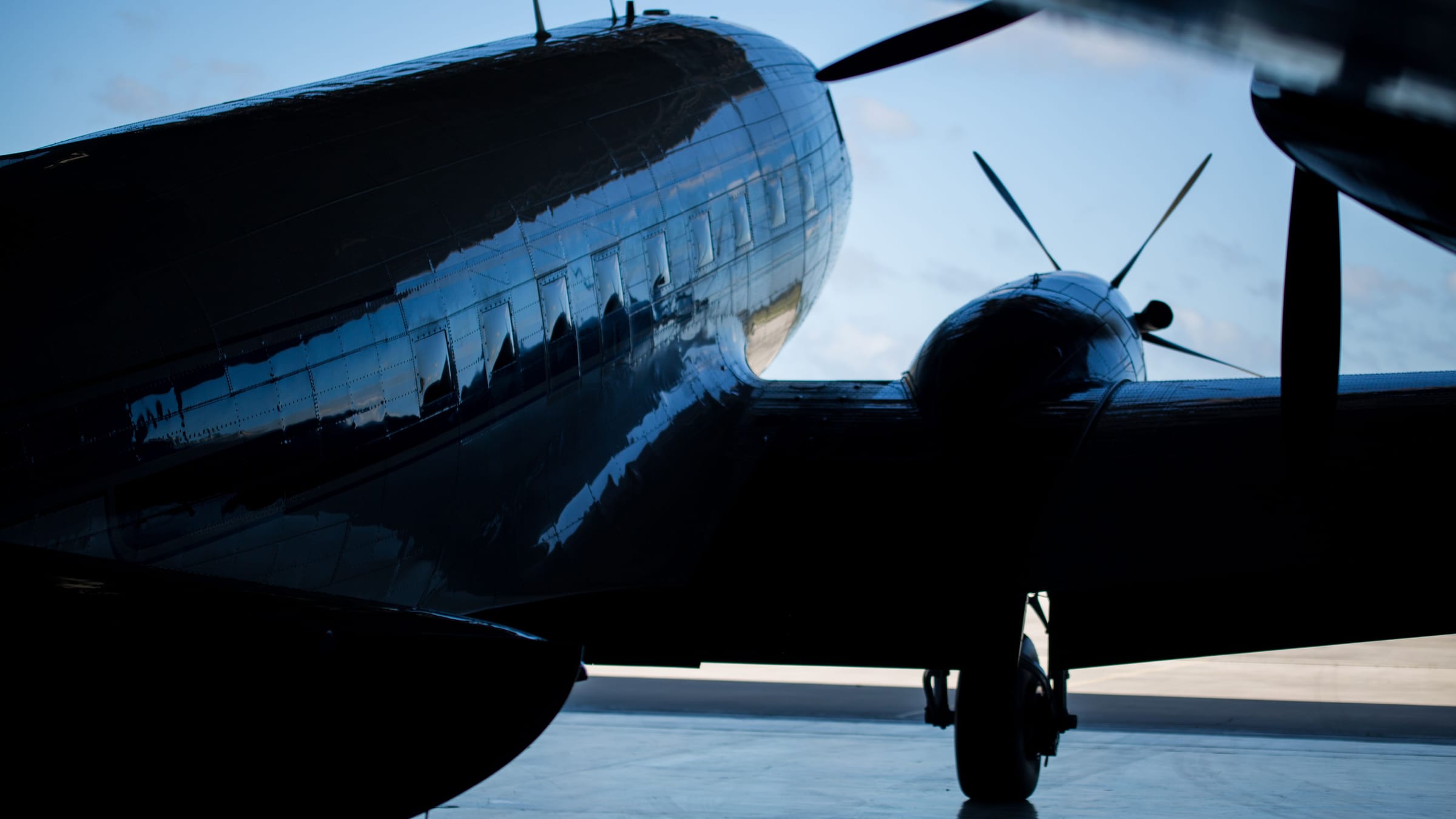 Polished fuselage and propeller engine of a vintage twin-engine aircraft in silhouette at a hangar door, with a blue evening sky beyond.